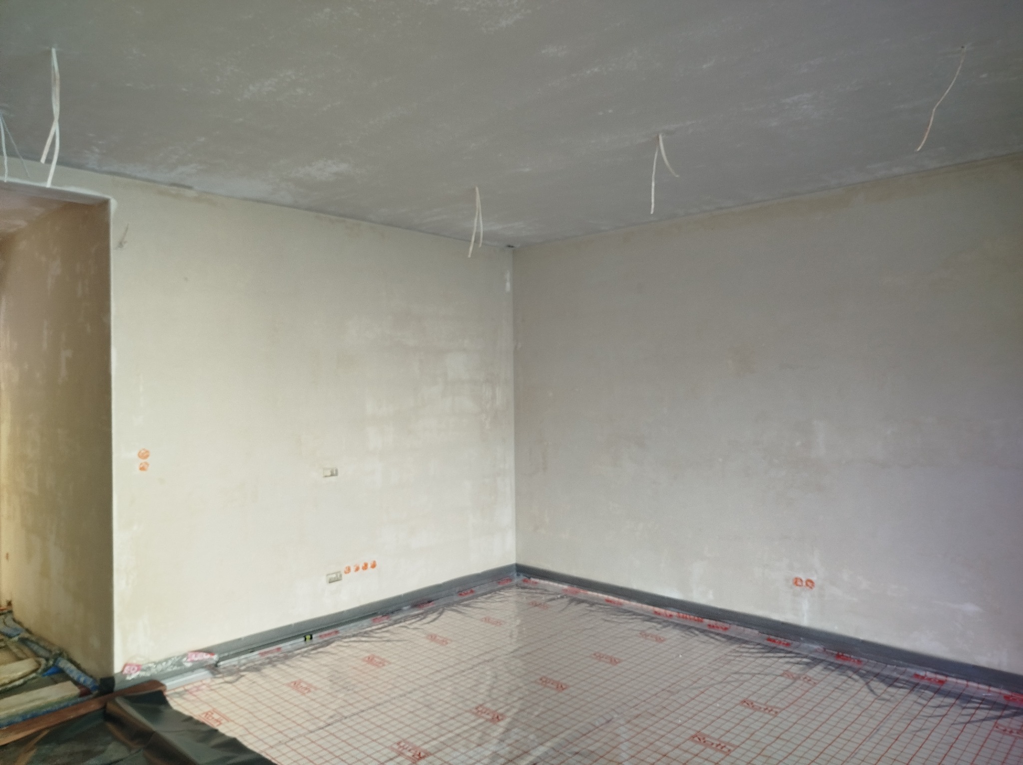 Wnętrze pokoju po nałożeniu świeżych, jasnych tynków na ścianach i suficie, podłoga zabezpieczona folią z siatką w kratkę, widoczne wystające kable elektryczne.