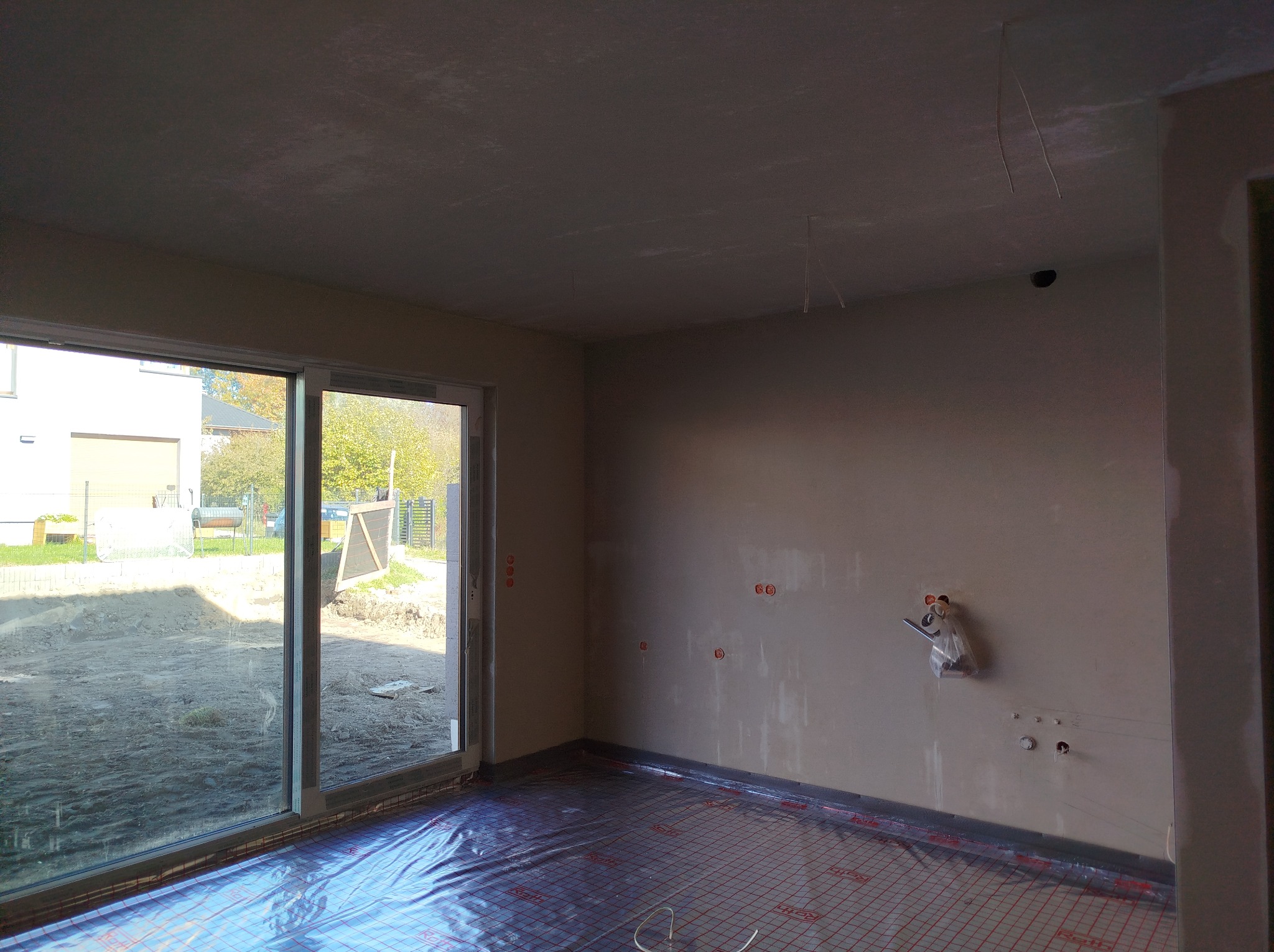 Wnętrze w trakcie prac tynkarskich maszynowych: surowy tynk na ścianach i suficie, zabezpieczona folią instalacja ogrzewania podłogowego, widoczne punkty elektryczne i hydrauliczne.