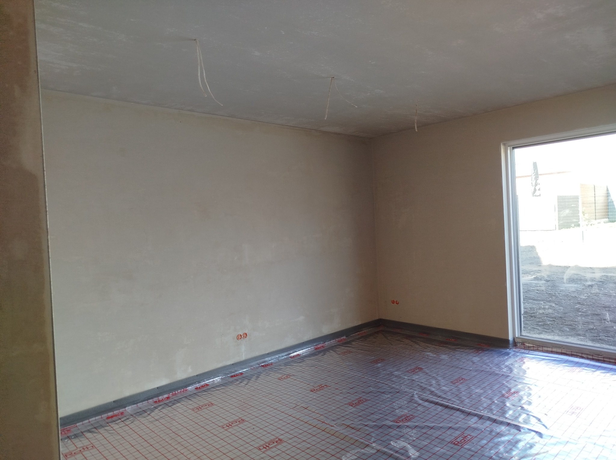 Świeżo otynkowane ściany w pomieszczeniu z folią grzewczą na podłodze, widoczne wystające kable z sufitu, przygotowane do dalszych prac wykończeniowych.