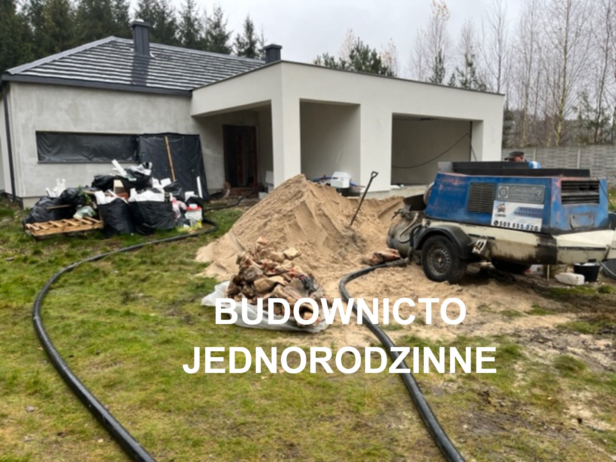 Budowa domu jednorodzinnego: widok na plac budowy z hałdą piasku, workami z odpadami budowlanymi i maszyną do wylewania posadzek, na tle surowego budynku z szarą dachówką.