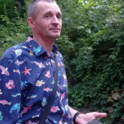 Mężczyzna w koszuli w ryby, stojący na tle zieleni w Ożarowie Mazowieckim, trzymający telefon w dłoni.