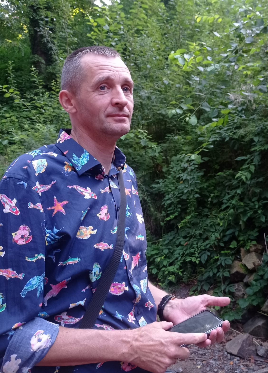 Mężczyzna w koszuli w ryby, stojący na tle zieleni w Ożarowie Mazowieckim, trzymający telefon w dłoni.