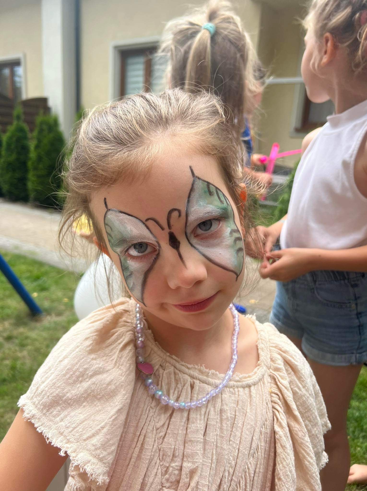 Dziewczynka z pomalowaną twarzą w motyla, nosząca naszyjnik z koralików, na tle zielonego trawnika i innych dzieci.