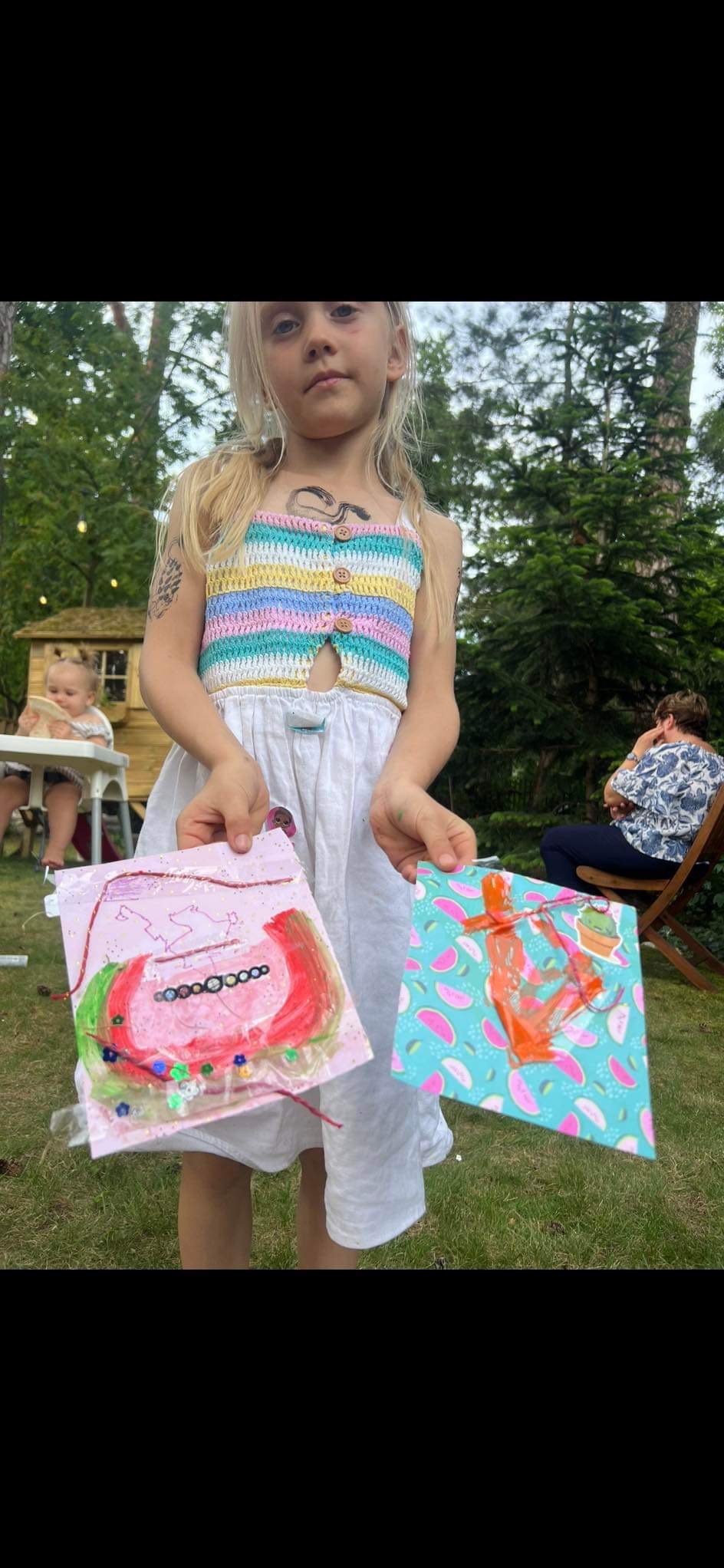 Dziewczynka w kolorowym topie prezentuje dwie prace plastyczne wykonane farbami i ozdobami, z widocznymi elementami zieleni w tle.