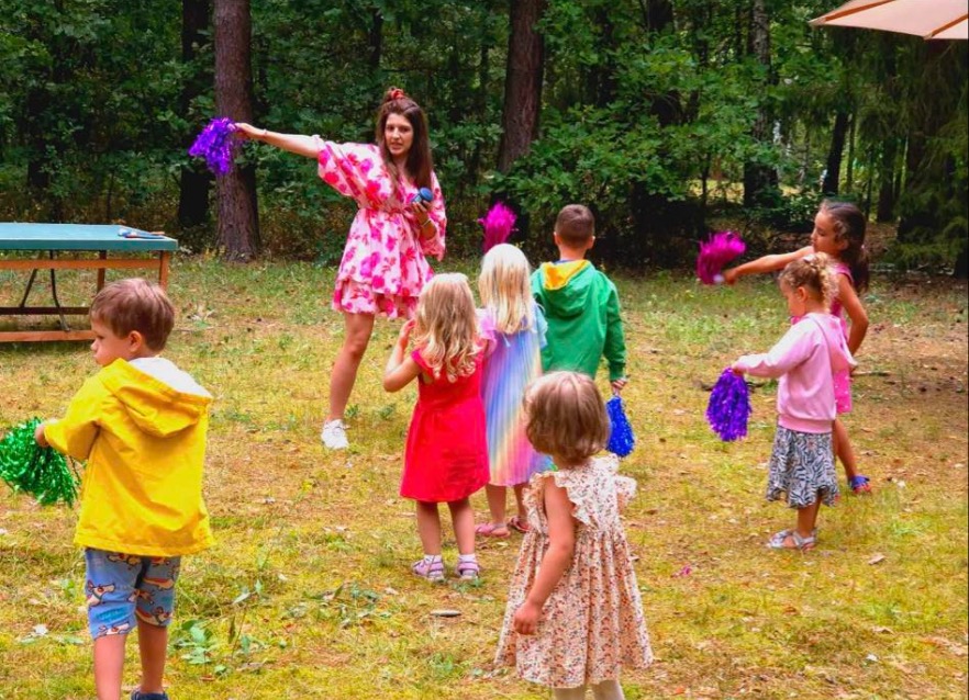 Kobieta w kwiecistej sukience prowadzi grupę dzieci z pomponami na leśnej polanie.