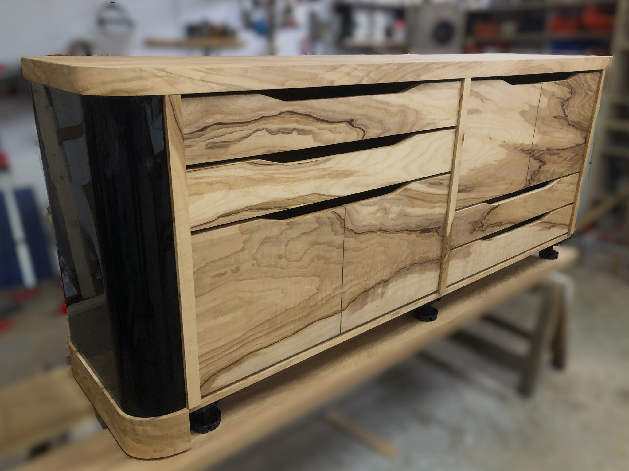 Komoda z forniru oliwkowego z lakierowanymi na wysoki połysk bokami, widoczna w warsztacie na drewnianym stole, z charakterystycznymi frezowanymi uchwytami szuflad.