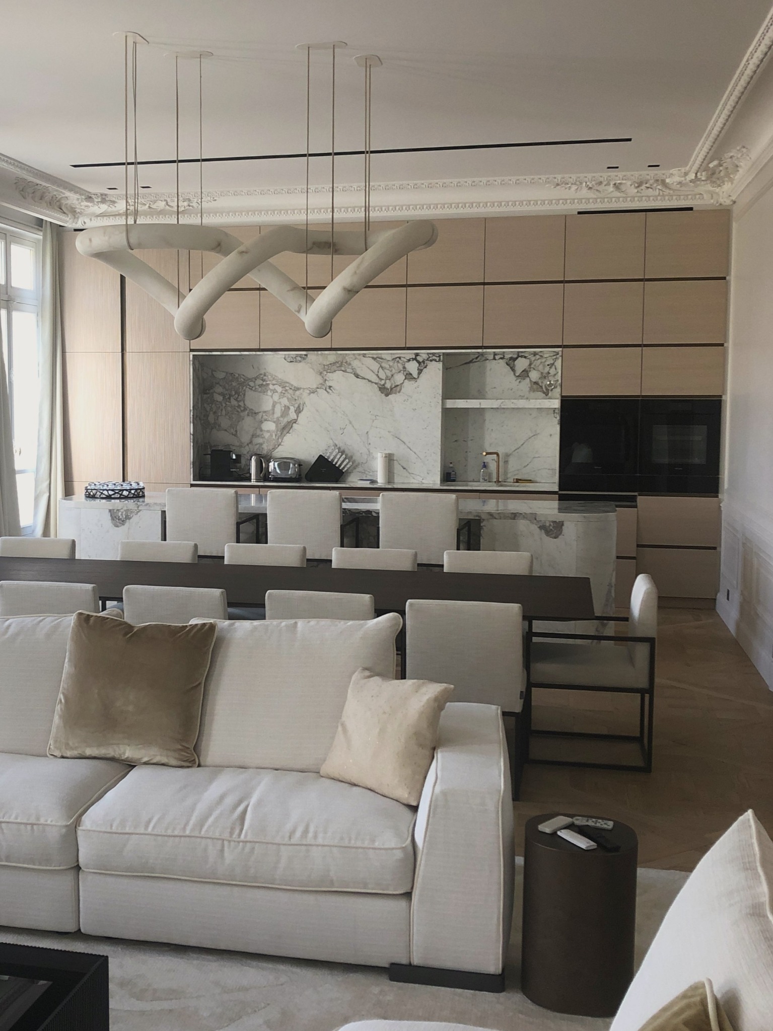 Nowoczesna kuchnia z zabudową z jasnego drewna i marmurowym blatem, wyspą i ścianą nad blatem, widziana z perspektywy salonu z białym narożnikiem i designerską lampą.