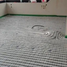 Białe rury ogrzewania podłogowego ułożone na siatce w surowym pomieszczeniu, zabezpieczone folią przy ścianach.