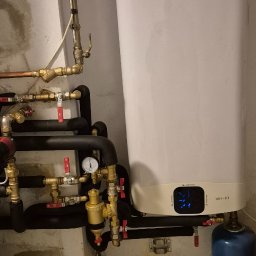 Firma Handlowo Usługowa WZOREK S.C - Biały, wiszący bojler elektryczny Ariston z widocznymi przyłączami hydraulicznymi i gazowymi, manometrem oraz niebieską butlą gazową w piwnicy z wilgotnymi ścianami i podłogą wyłożoną płytkami.
