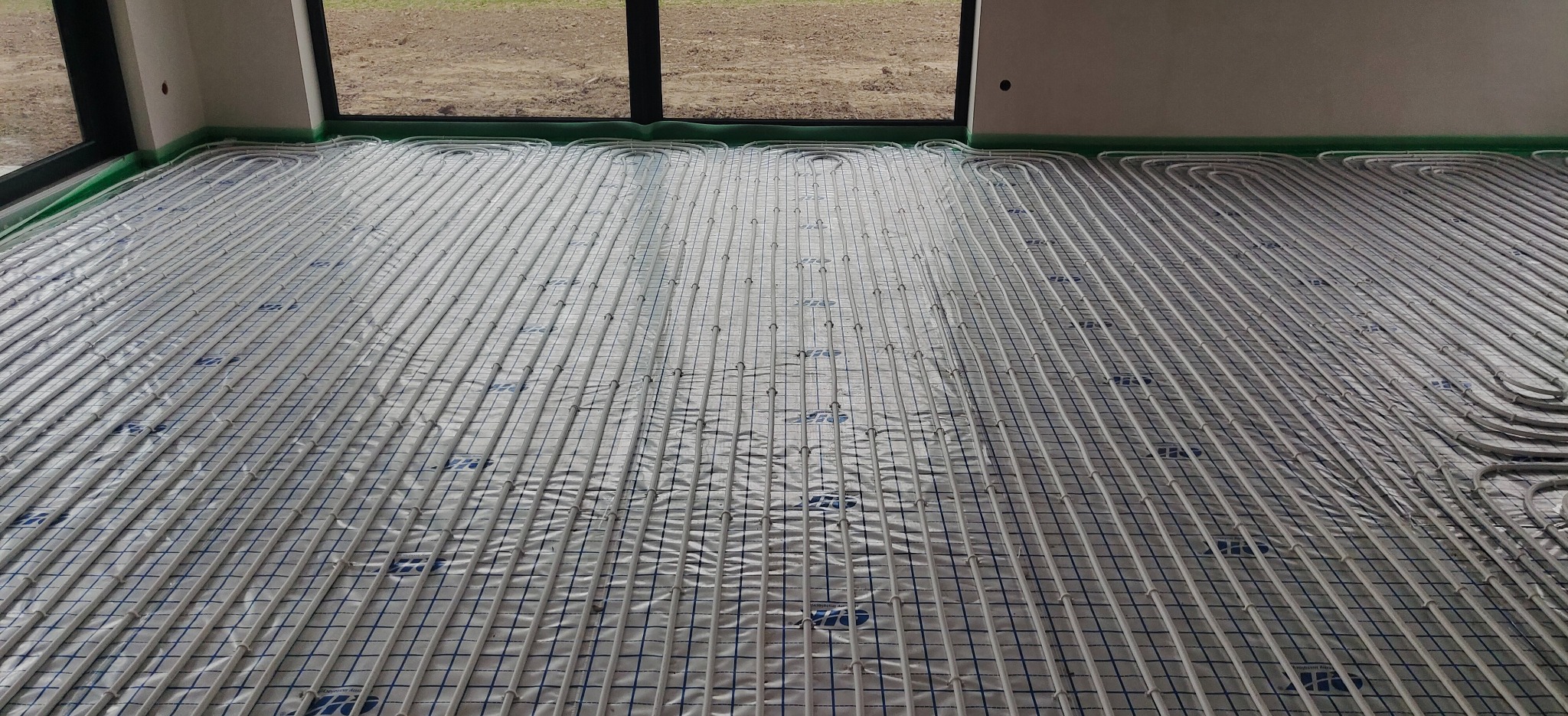 Rozłożone rury ogrzewania podłogowego na folii z nadrukowaną siatką, widoczne okno i fragment ściany, przygotowanie do wylewki.