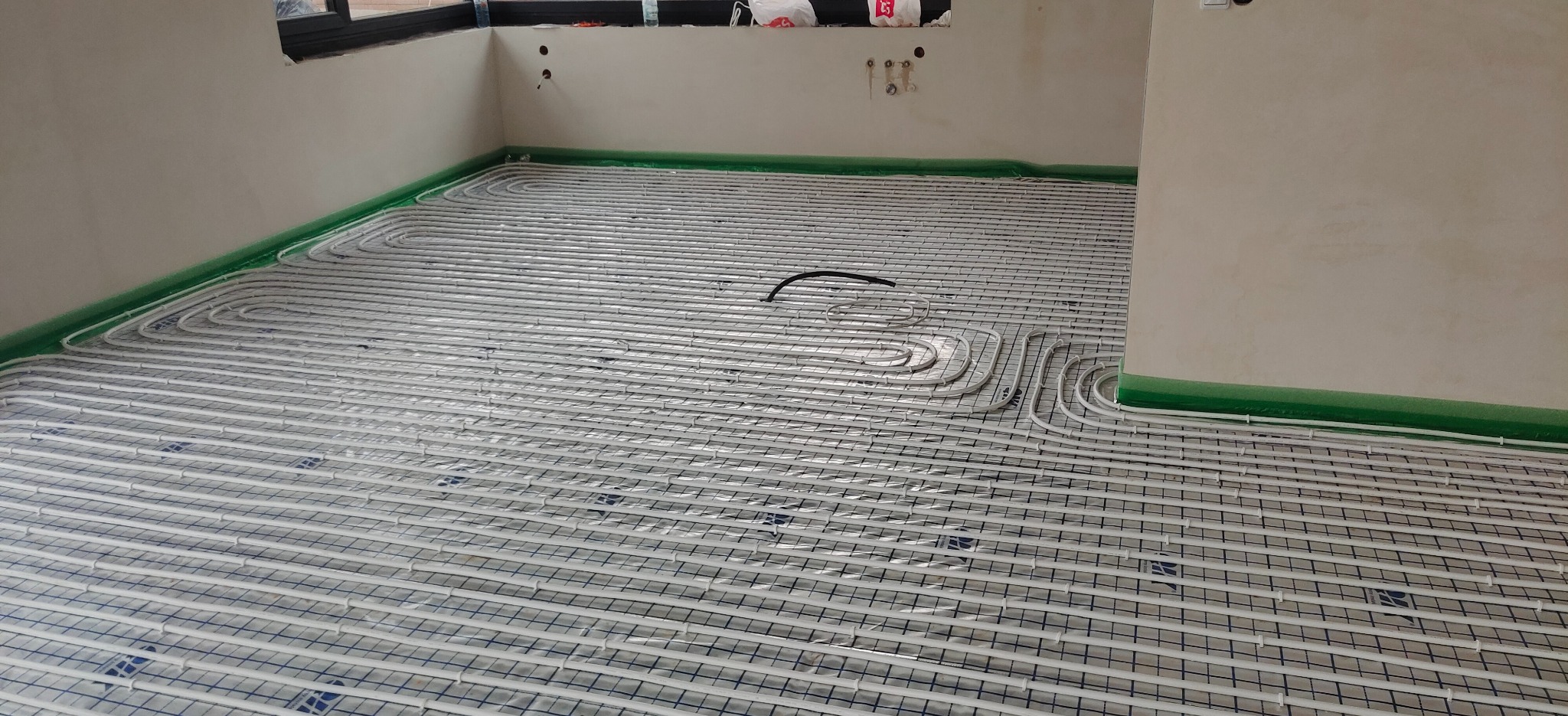 Białe rury ogrzewania podłogowego ułożone na siatce w surowym pomieszczeniu, zabezpieczone folią przy ścianach.