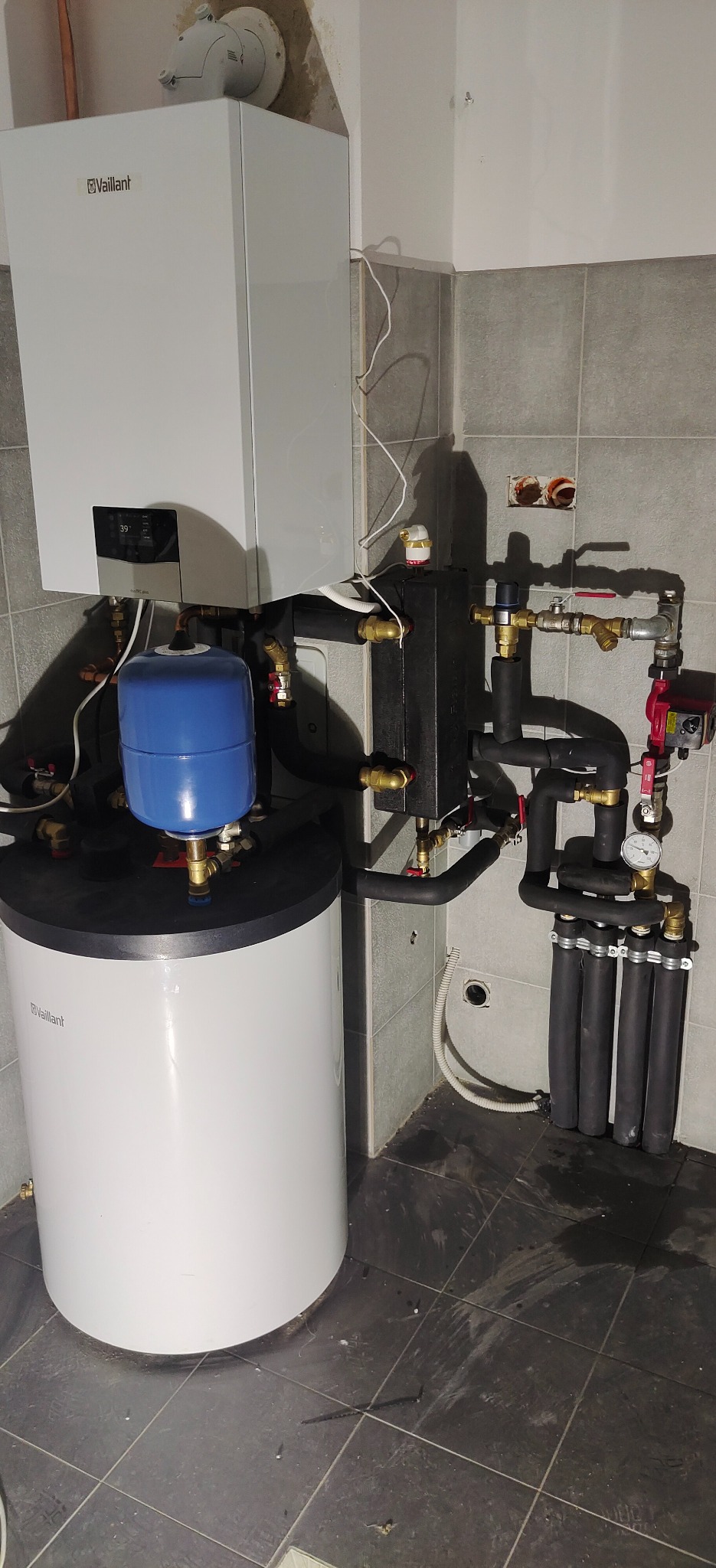 Biały piec gazowy Vaillant z podłączonym zasobnikiem wody i elementami instalacji grzewczej na tle szarej, kafelkowanej ściany w pomieszczeniu. Widoczne rury, zawory i czarny rozdzielacz.