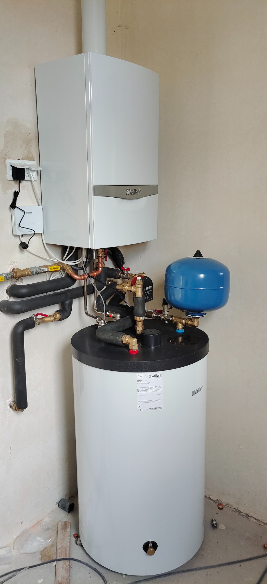 Biały piec gazowy kondensacyjny Vaillant zamontowany na białym zasobniku ciepłej wody użytkowej, z widocznymi rurami miedzianymi i czarnymi izolowanymi, oraz niebieskim naczyniem przeponowym,...