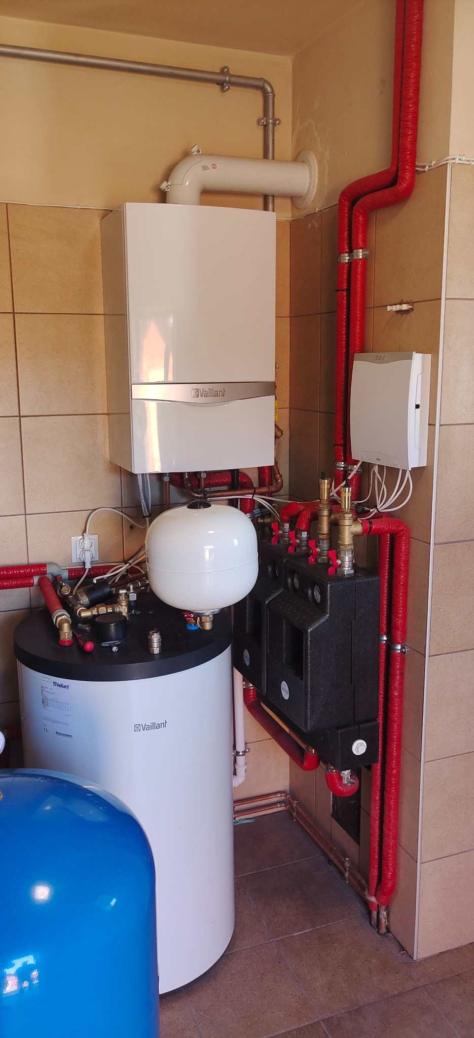 Nowoczesna kotłownia z białym piecem gazowym Vaillant, zasobnikiem ciepłej wody, rozdzielaczem grzewczym i rurami w czerwonej izolacji, zamontowana w pomieszczeniu wyłożonym beżowymi płytkami.