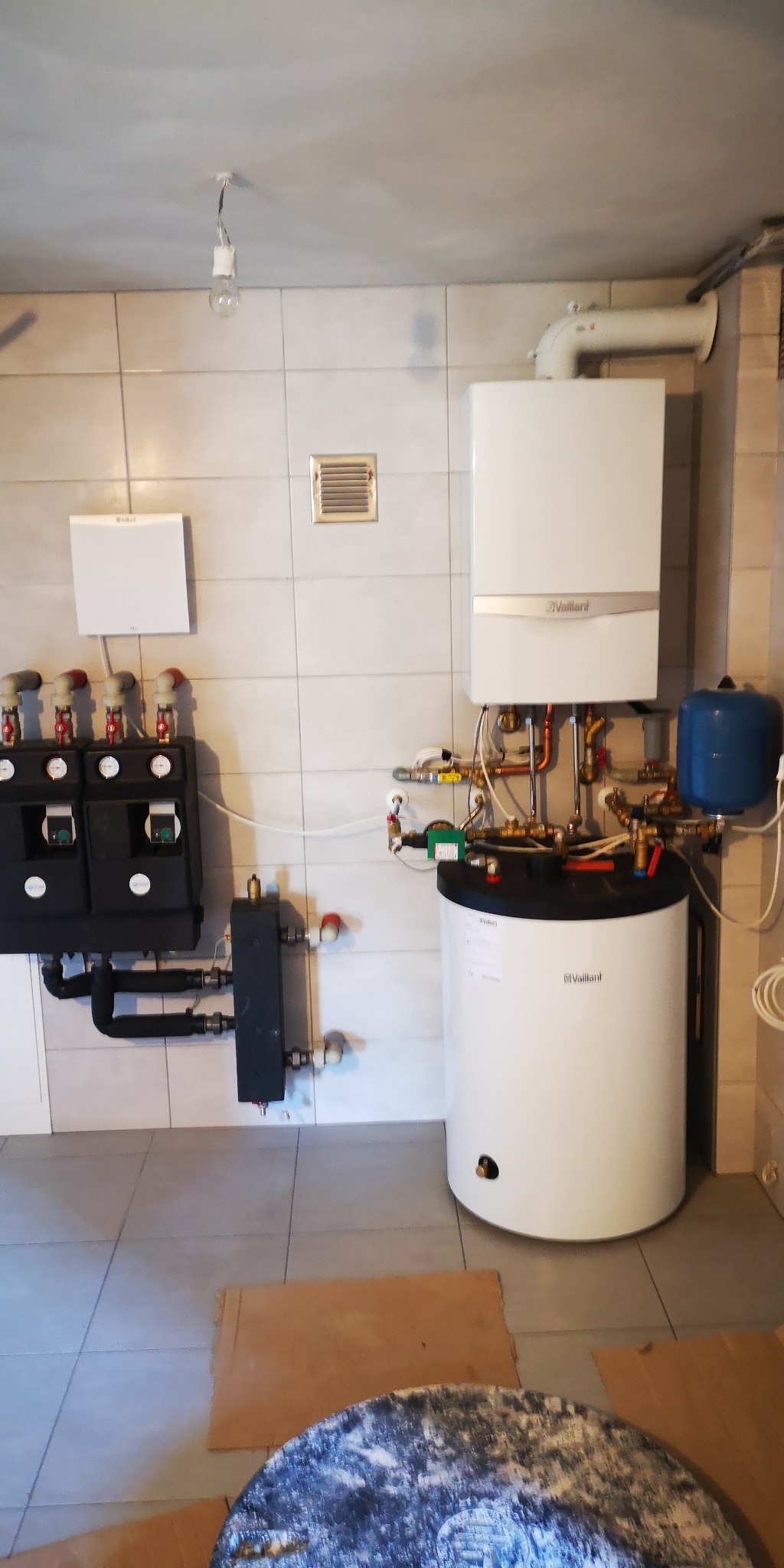 Biały, wiszący piec gazowy Vaillant z podłączonymi rurami miedzianymi i niebieskim zbiornikiem wyrównawczym, zamontowany na kafelkowanej ścianie, obok rozdzielaczy ogrzewania podłogowego,...