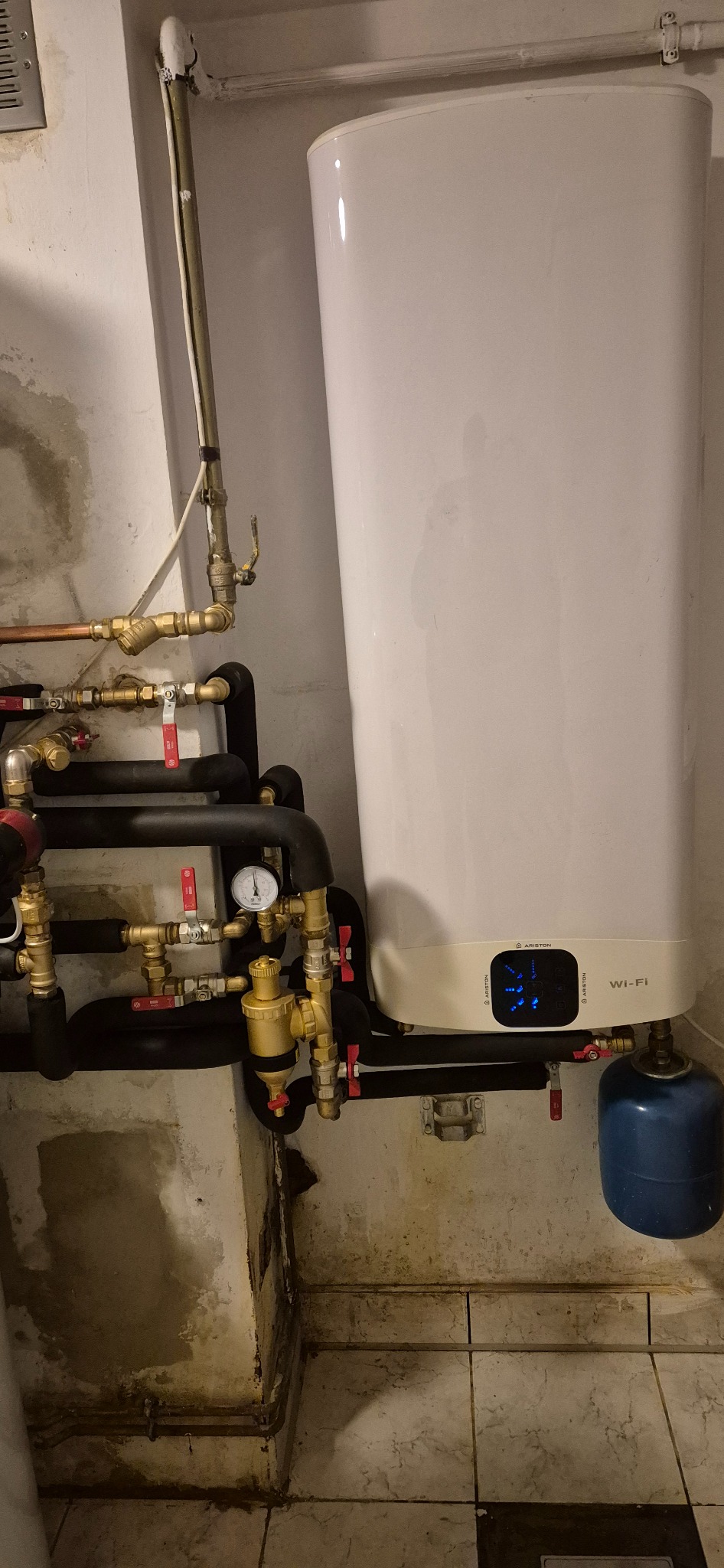 Biały, wiszący bojler elektryczny Ariston z widocznymi przyłączami hydraulicznymi i gazowymi, manometrem oraz niebieską butlą gazową w piwnicy z wilgotnymi ścianami i podłogą wyłożoną płytkami.