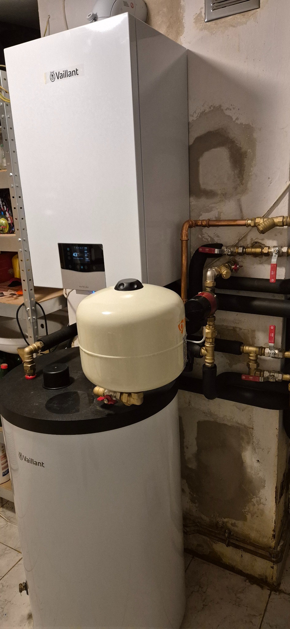 Biały piec gazowy Vaillant z wyświetlaczem, pod nim biały zasobnik wody z beżowym naczyniem przeponowym, widoczne miedziane rury i zawory na tle ściany z zaciekami.