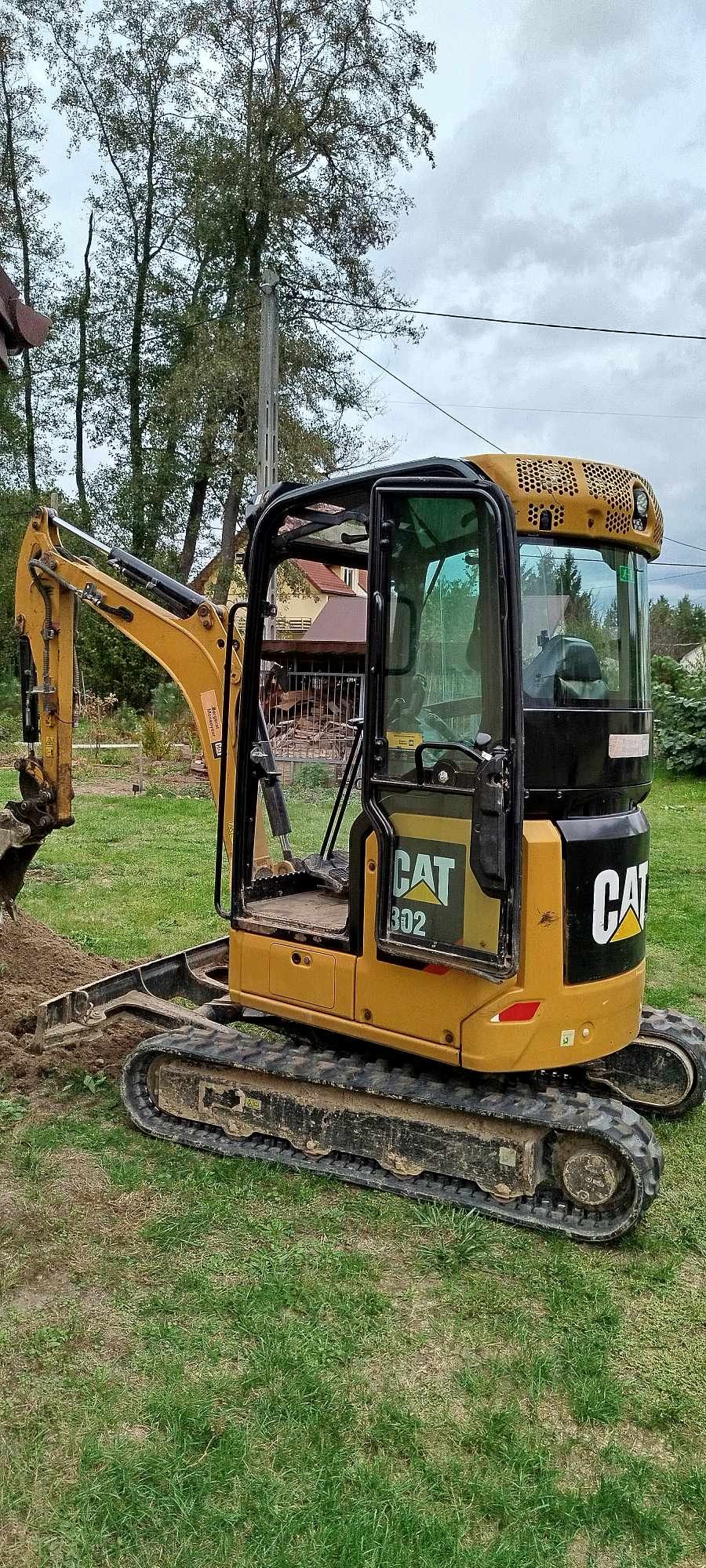 Żółta minikoparka CAT 302 w trakcie prac ziemnych na trawiastym terenie, widoczne ślady świeżo przekopanej ziemi, w tle dom jednorodzinny i drzewa.