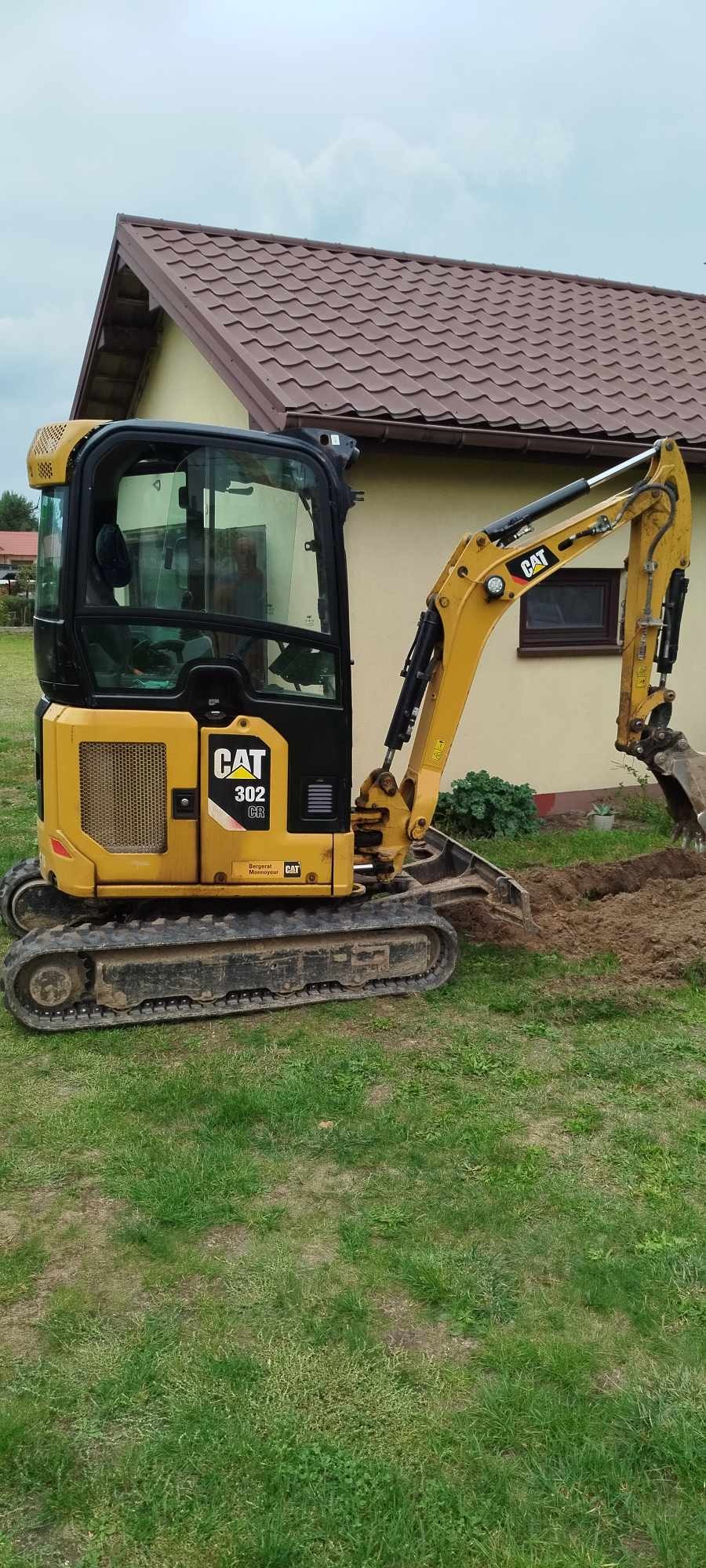 Żółta mini koparka CAT 302 CR podczas wykonywania wykopu w ziemi, z widocznym budynkiem mieszkalnym w tle.