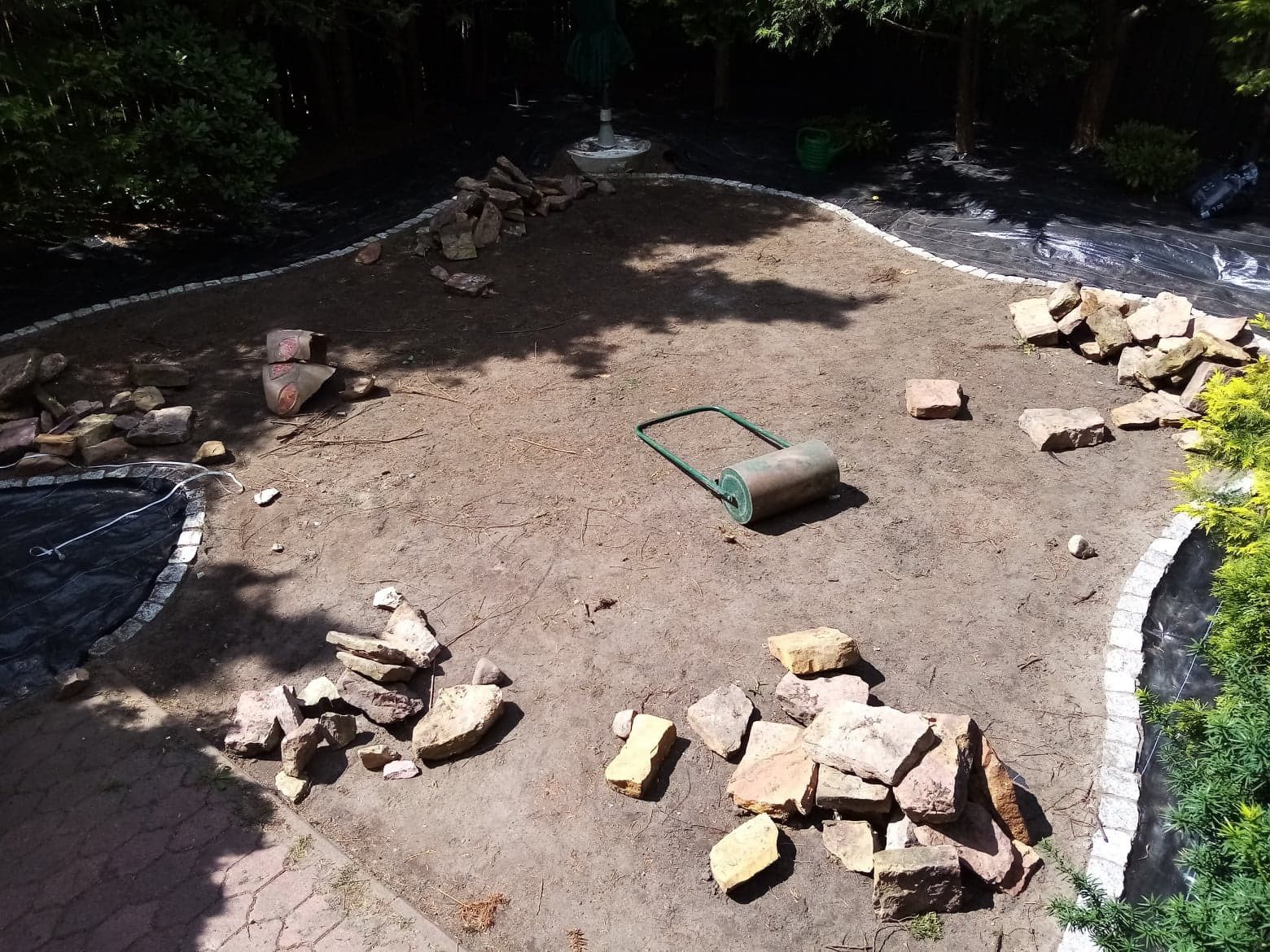 Przygotowany teren pod trawnik z rolki: rozłożona agrowłóknina, ułożone obrzeża z kostki i kamieni, ziemia wyrównana walcem ogrodowym, stosy kamieni dekoracyjnych.