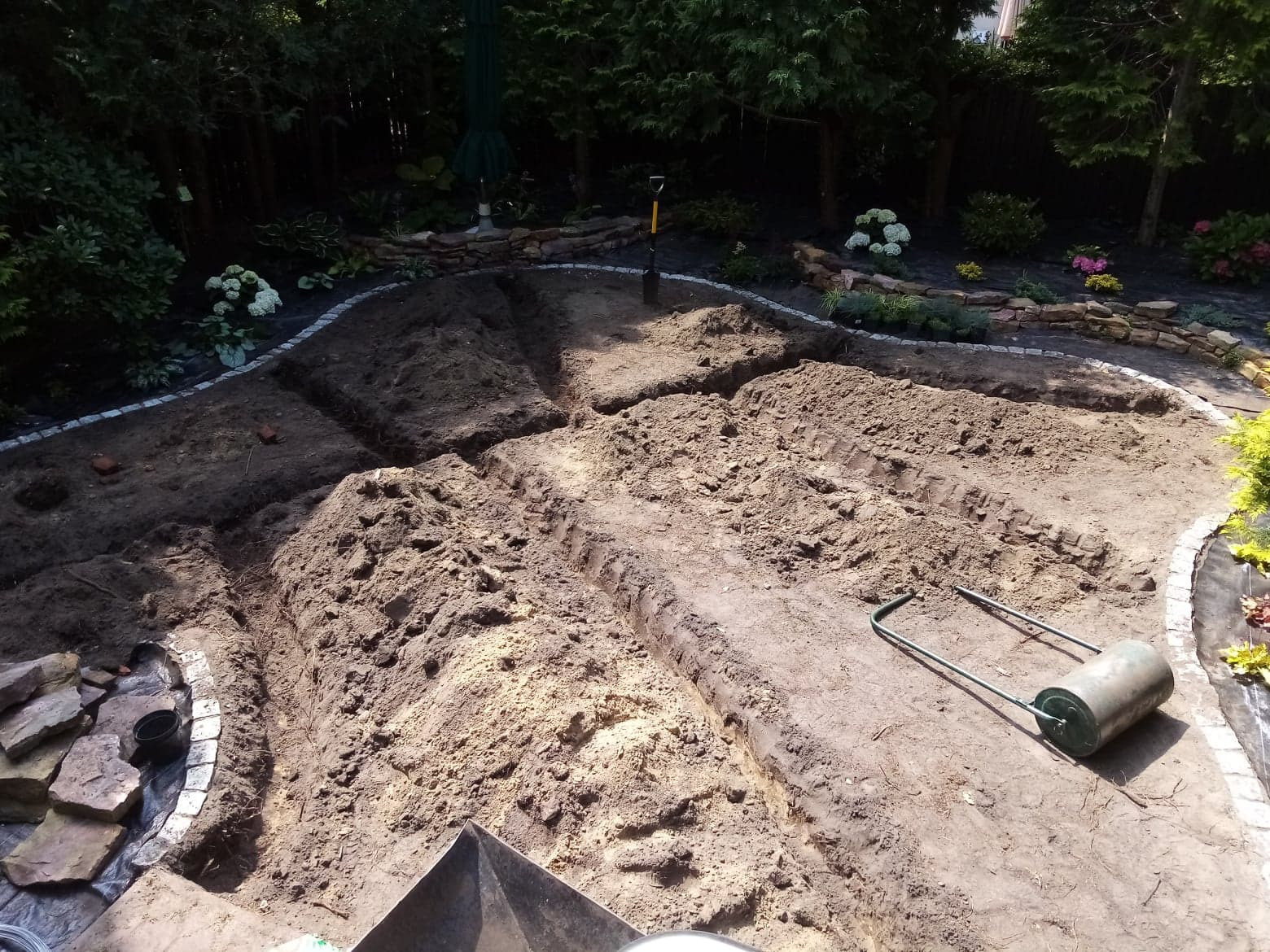 Przygotowany teren pod przyszły ogród z wykopanymi rowami, widoczna ubijarka do ziemi, łopata i kamienne obrzeża.