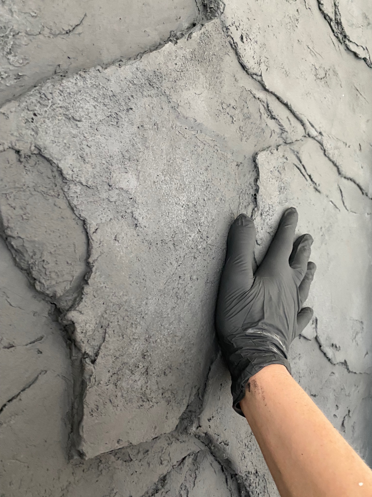 Tekstura ściany imitująca kamień, pokryta szarą farbą, dłoń w czarnej rękawicy dotyka powierzchni.