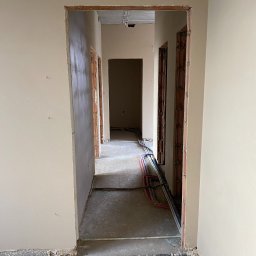 MSK-SYSTEM - Korytarz w trakcie prac wykończeniowych: ściany pokryte świeżym, jasnym tynkiem, widoczne instalacje elektryczne i hydrauliczne prowadzone wzdłuż podłogi, surowy beton na podłodze, przygotowane...