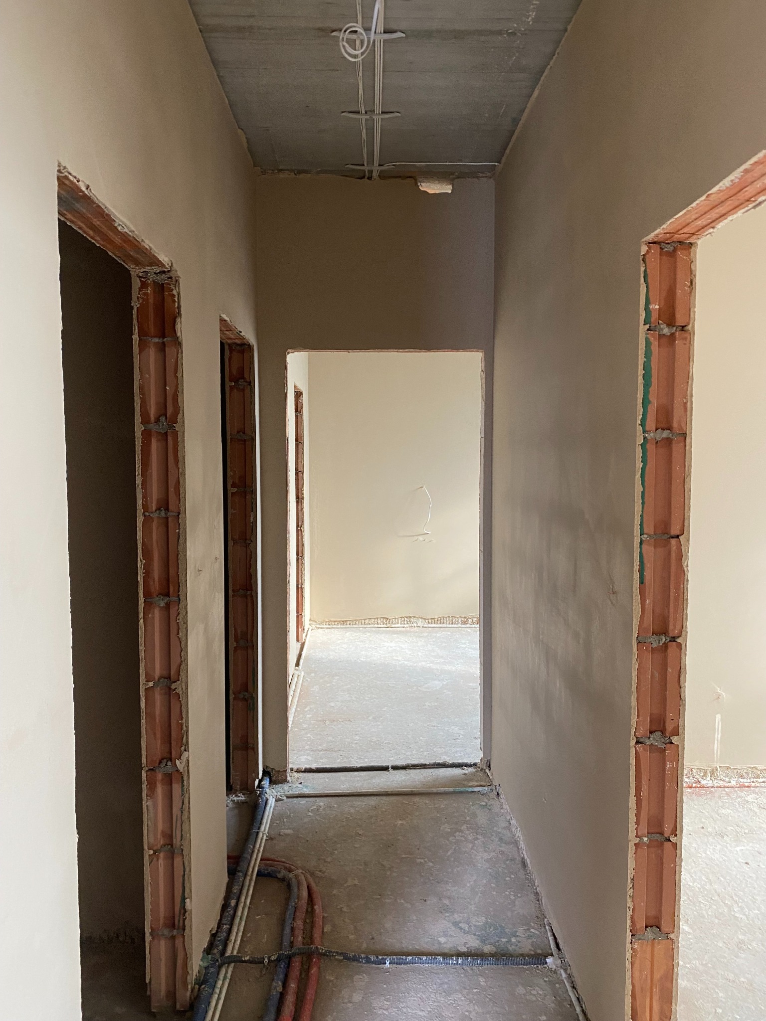 Korytarz w trakcie tynkowania maszynowego, widoczne surowe ściany z cegły wokół otworów drzwiowych i rury instalacyjne na podłodze.