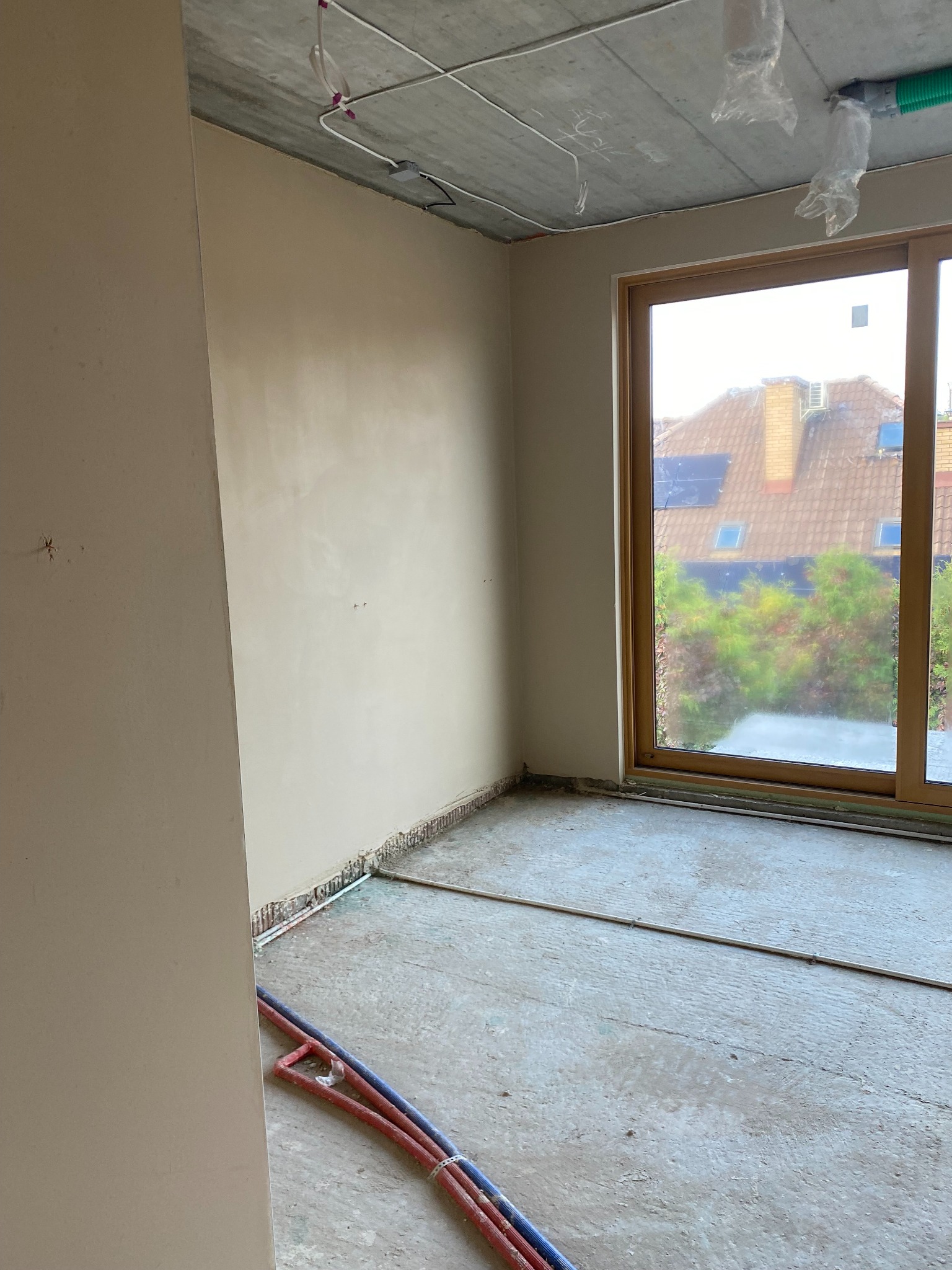 Pokój w stanie deweloperskim po nałożeniu gładzi, widoczne przewody instalacji podłogowej, surowy betonowy sufit z instalacjami podwieszonymi, okno z widokiem na dom z panelami słonecznymi.