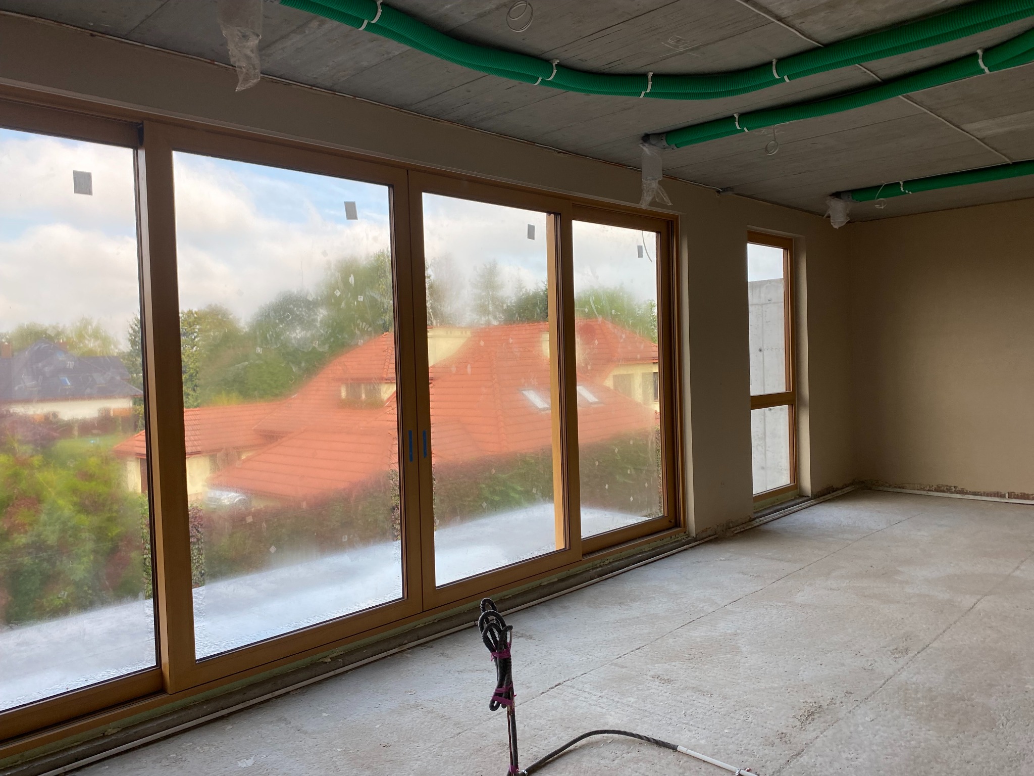 Wnętrze w stanie deweloperskim z dużymi przesuwnymi oknami, widoczne zielone rury wentylacyjne pod sufitem i sprzęt do natrysku tynku na podłodze.