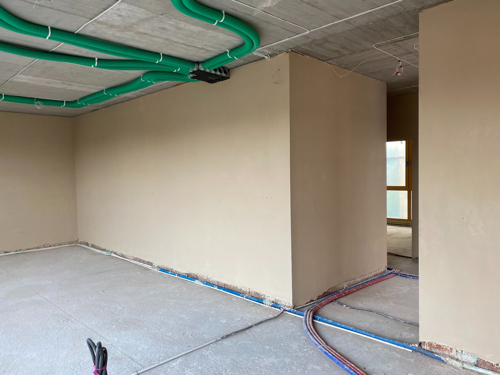 Wnętrze w trakcie prac wykończeniowych: ściany po nałożeniu gładzi gipsowej, widoczne instalacje elektryczne w zielonych rurach na suficie oraz rury instalacyjne wzdłuż podłogi, surowy betonowy sufit.