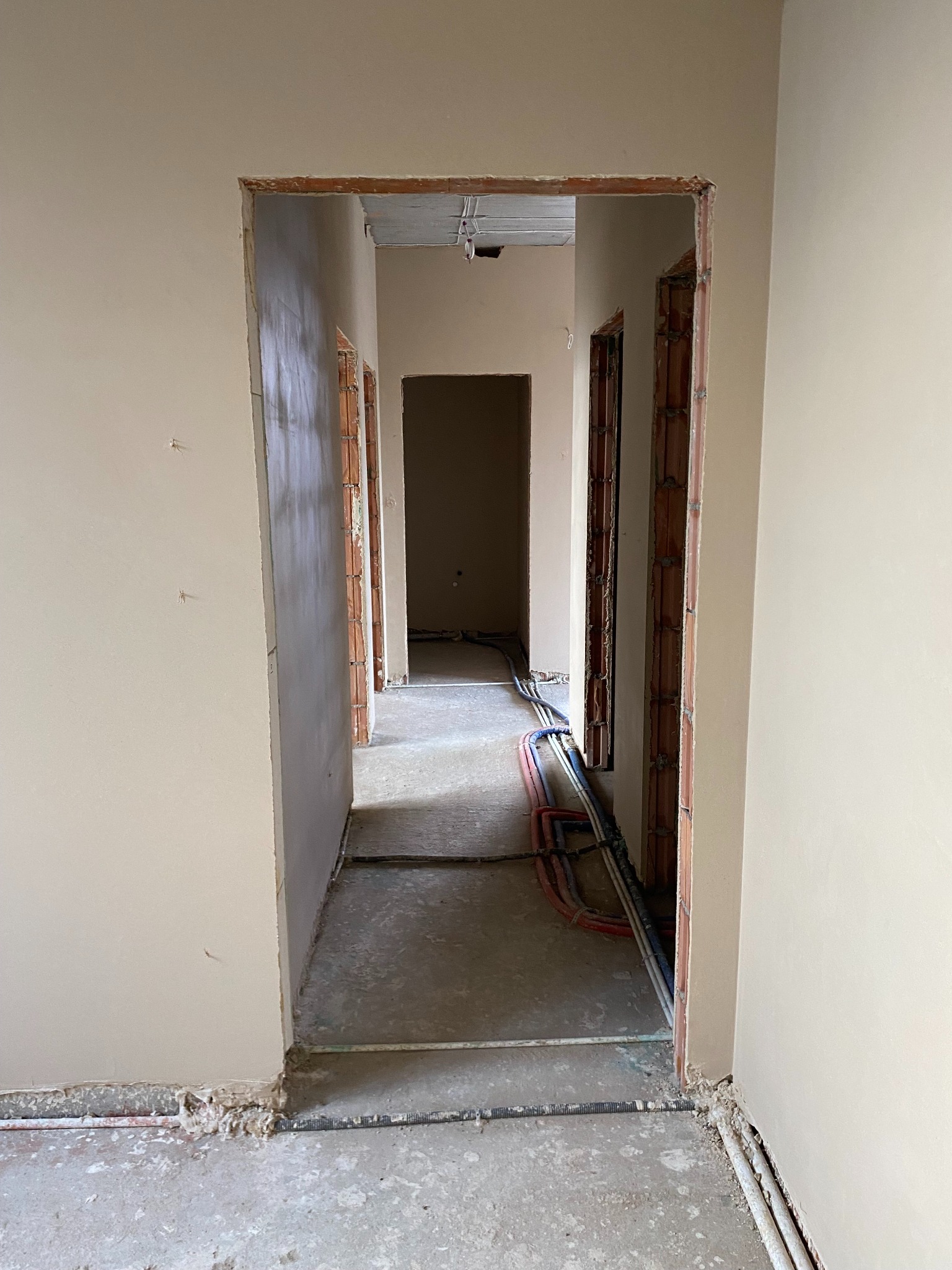 Korytarz w trakcie prac wykończeniowych: ściany pokryte świeżym, jasnym tynkiem, widoczne instalacje elektryczne i hydrauliczne prowadzone wzdłuż podłogi, surowy beton na podłodze, przygotowane...