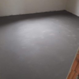 Wylewki maszynowe jastrychowe - Świeżo wylana, gładka, szara podłoga w pomieszczeniu z białymi ścianami i oknem, widoczny fragment muru.