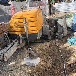 Wylewki maszynowe jastrychowe - Żółta maszyna Putzmeister na placu budowy przed budynkiem w stylu góralskim, z widocznymi elementami konstrukcyjnymi i gruzem.