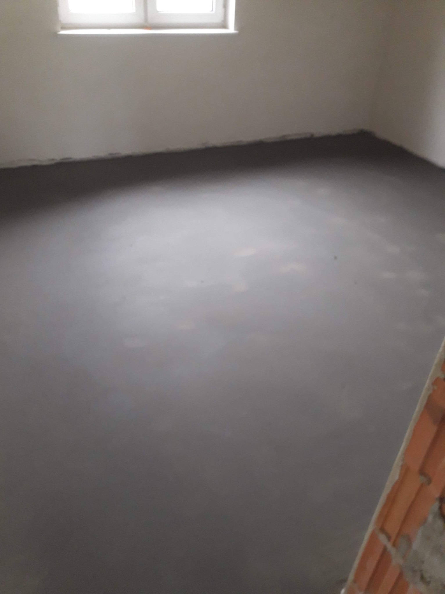 Świeżo wylana, gładka, szara podłoga w pomieszczeniu z białymi ścianami i oknem, widoczny fragment muru.