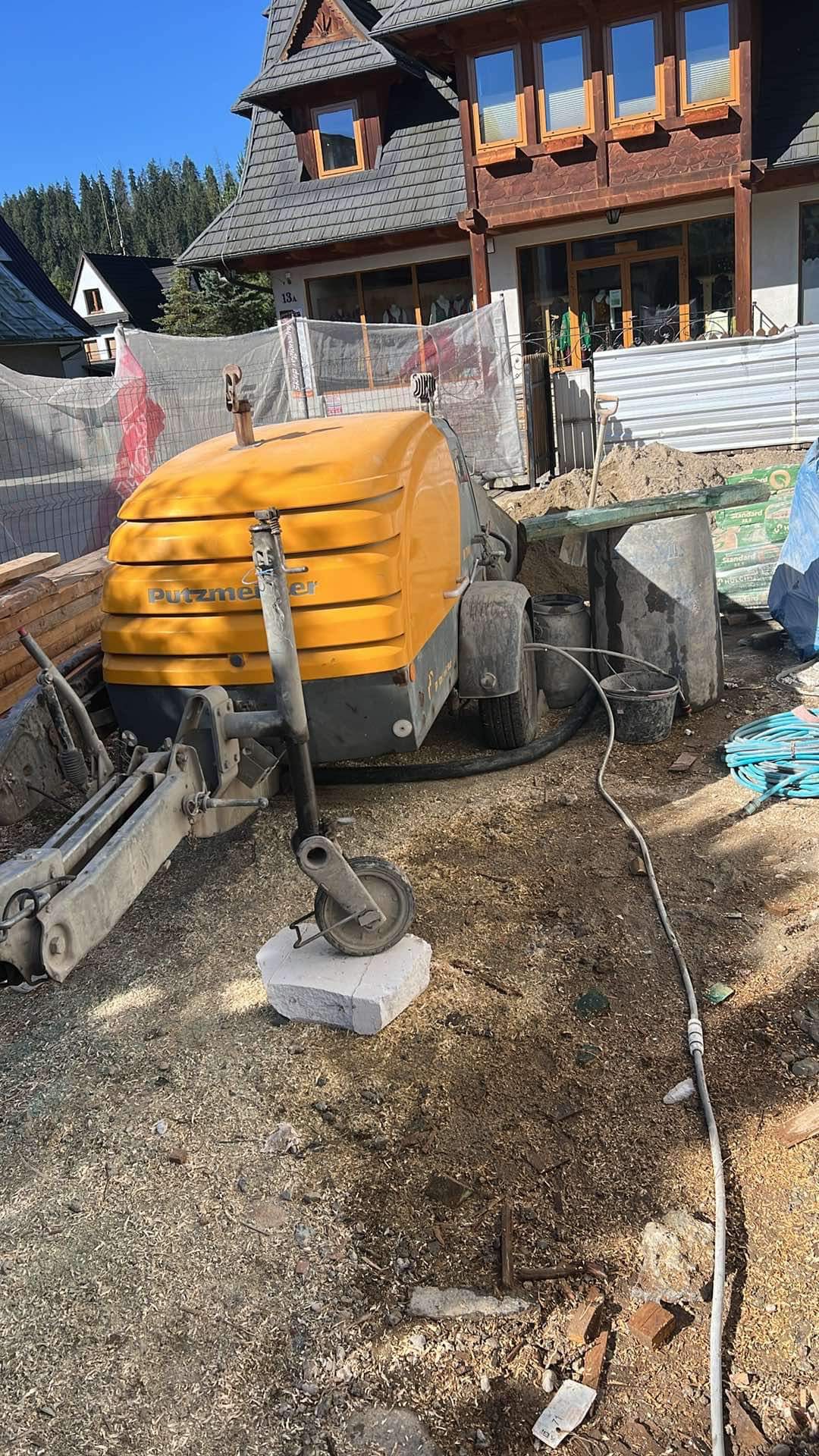 Żółta maszyna Putzmeister na placu budowy przed budynkiem w stylu góralskim, z widocznymi elementami konstrukcyjnymi i gruzem.