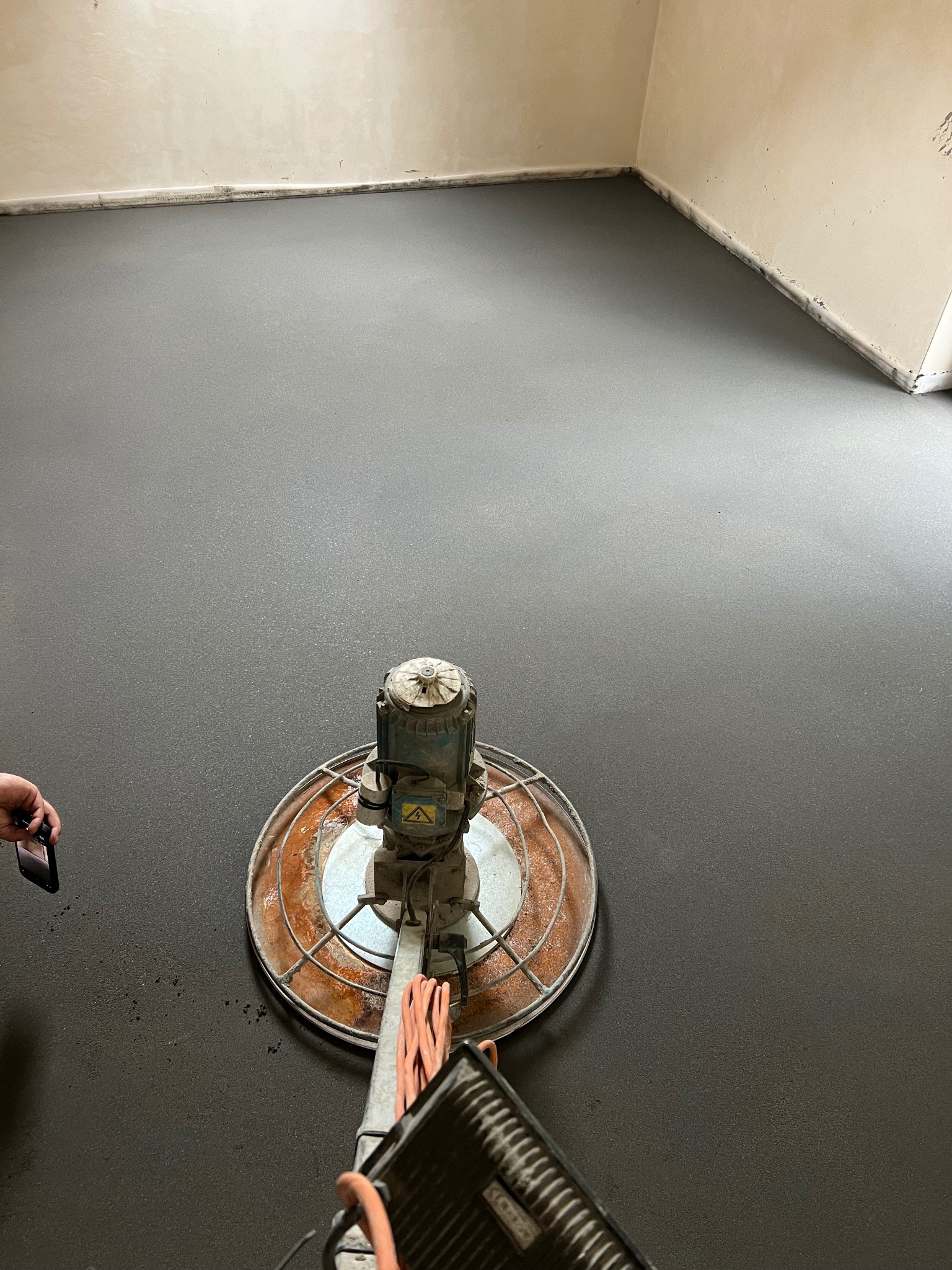 Szlifowanie świeżej, ciemnoszarej posadzki jastrychowej maszyną z metalową osłoną, widoczny fragment ręki z telefonem.