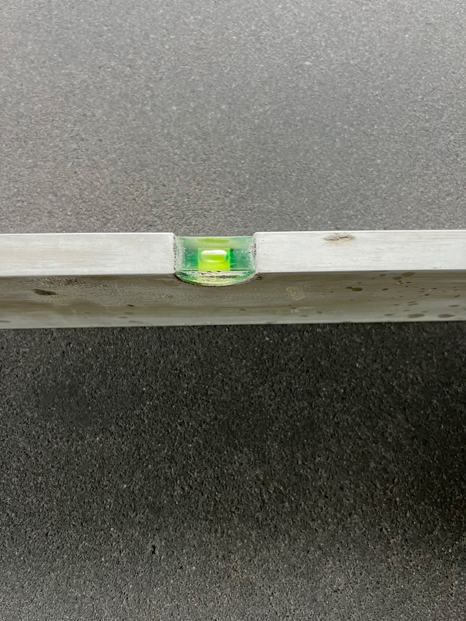 Poziomica aluminiowa z zielonym wskaźnikiem pęcherzykowym, leżąca na ciemnej, szorstkiej powierzchni. Widoczne drobne zabrudzenia i zarysowania na poziomicy.