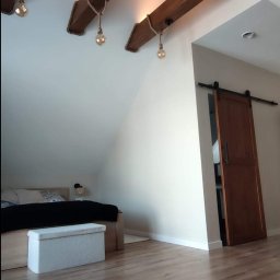 Spec Remonty - Adaptacja poddasza: drewniane belki sufitowe z żarówkami na sznurach, przesuwne drzwi do garderoby, jasna podłoga z paneli i skosy sufitowe nad łóżkiem.