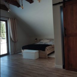 Spec Remonty - Pokój na poddaszu z drewnianą podłogą, belkami stropowymi z oświetleniem sznurkowym i przesuwnymi drzwiami do sypialni.