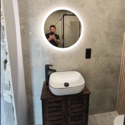 Spec Remonty - Umywalka nablatowa w łazience z okrągłym lustrem LED, widoczne szare płytki, czarna bateria i fragment ściany przed wykończeniem.