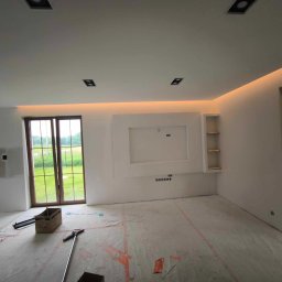 Spec Remonty - Wnętrze w trakcie wykończenia: białe ściany z podświetleniem LED, przygotowane podłoże pod panele, widoczne narzędzia i karton po płytkach podłogowych.