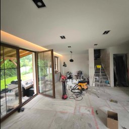 Spec Remonty - Wnętrze w trakcie prac wykończeniowych: otwarte przesuwne drzwi balkonowe z widokiem na zieleń, przygotowane podłoże, drabina, lampa wisząca, sprzęt budowlany i zakryte folią schody.