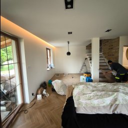 Spec Remonty - Montaż parkietu w jodełkę w jasnym wnętrzu z oknami i ceglaną ścianą, widoczny pracownik układający panele i drabina w tle.