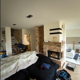 Spec Remonty - Remontowane wnętrze domu: pracownik układa panele pod schodami, widoczny kominek z cegły, zabezpieczone meble i narzędzia pracy.