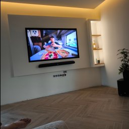 Spec Remonty - Telewizor wiszący na ścianie z podświetleniem LED, półka wnękowa z dekoracjami, parkiet ułożony w jodełkę, widoczny fragment stopy osoby siedzącej na pufie.