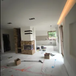 Przestronne wnętrze w trakcie prac wykończeniowych, z zabezpieczoną folią podłogą, ceglanym kominkiem z czarnymi akcentami, widocznymi narzędziami i materiałami budowlanymi oraz oświetleniem LED...