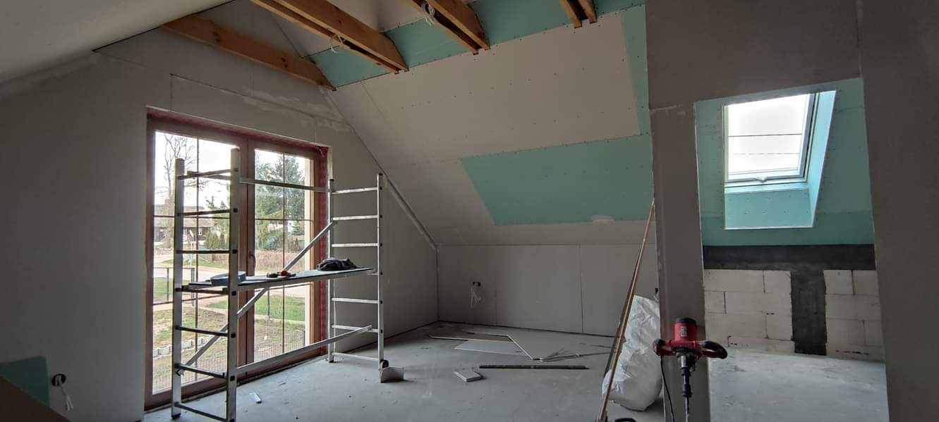 Poddasze w trakcie prac wykończeniowych: widoczne płyty gipsowo-kartonowe na skosach sufitu, okno dachowe, okno w szczycie, rusztowanie przy oknie, ściany przygotowane do dalszych prac, mieszadło...