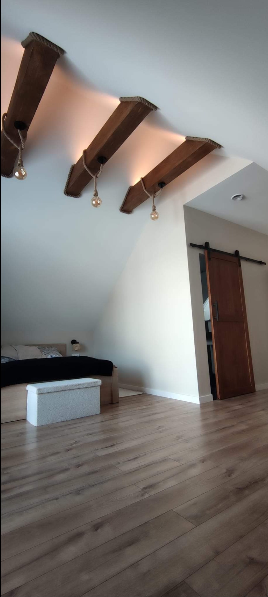 Adaptacja poddasza: drewniane belki sufitowe z żarówkami na sznurach, przesuwne drzwi do garderoby, jasna podłoga z paneli i skosy sufitowe nad łóżkiem.