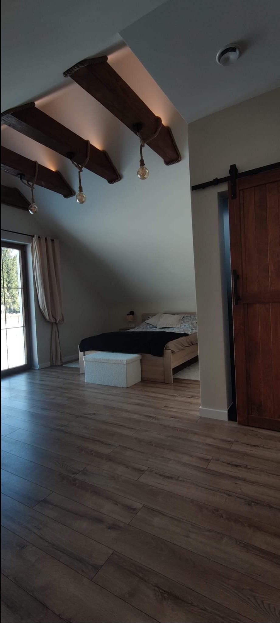 Pokój na poddaszu z drewnianą podłogą, belkami stropowymi z oświetleniem sznurkowym i przesuwnymi drzwiami do sypialni.